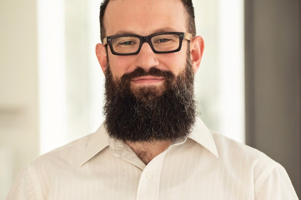 A man with dark hair, a beard, and glasses wearing a white button-up shirt is smiling at the camera.