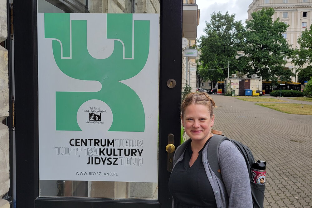 A smiling blonde woman standing next to a sign in Polish and Yiddish.