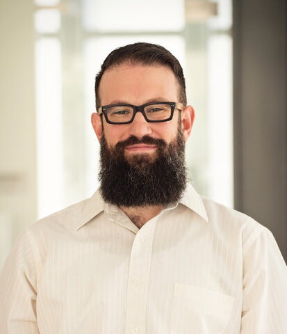 A man with dark hair, a beard, and glasses wearing a white button-up shirt is smiling at the camera.