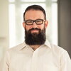 A man with dark hair, a beard, and glasses wearing a white button-up shirt is smiling at the camera.