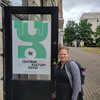 A smiling blonde woman standing next to a sign in Polish and Yiddish.