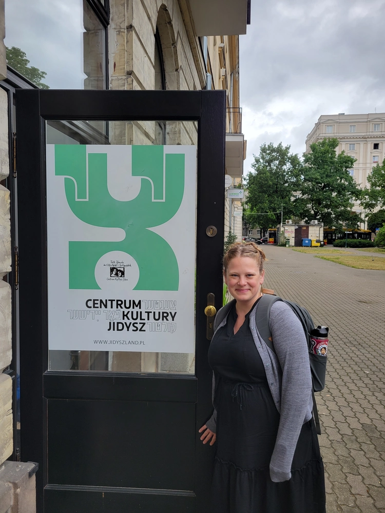 A smiling blonde woman standing next to a sign in Polish and Yiddish.